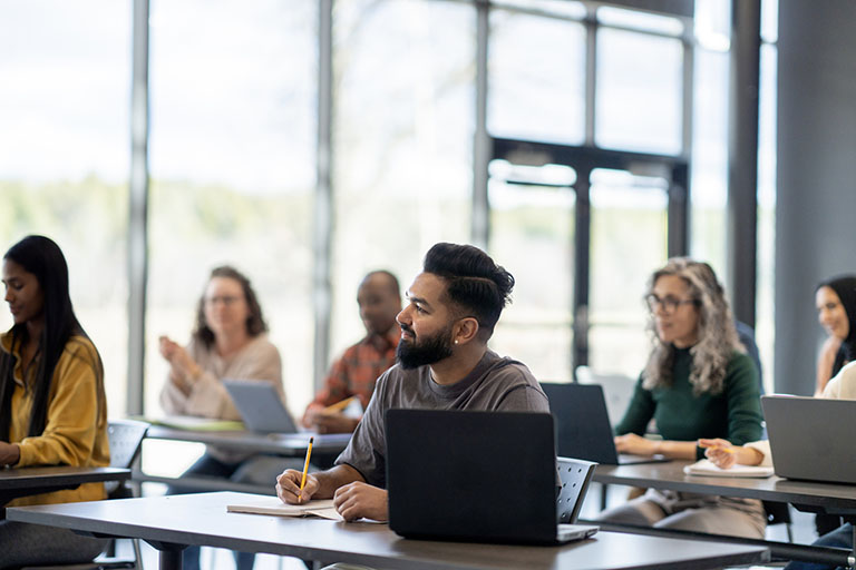 A large group of mature adults are seen sitting at desk with wither laptops and books open as they study during a continuing education class.